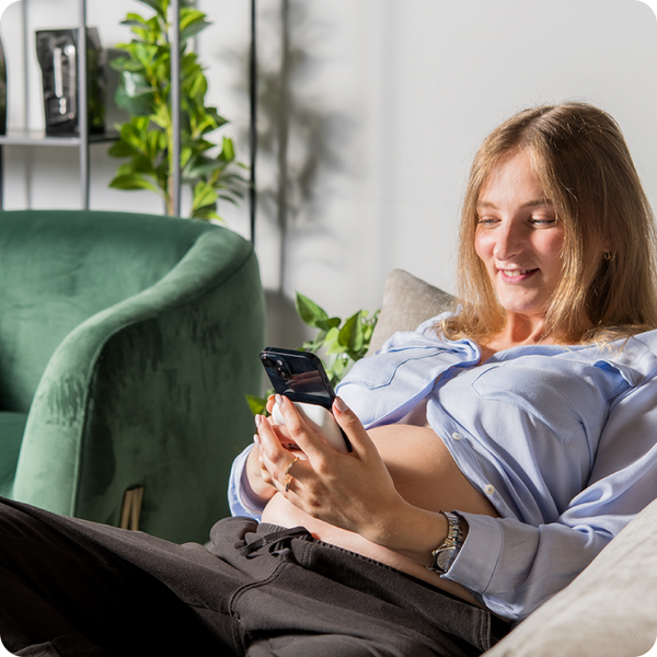Woman sitting on a couch using Pulsenmore home ultrasound device while scanning her belly  in a bright living room.